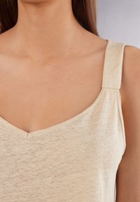 Světle béžový top na ramínka s širokými popruhy a designem ve tvaru V. Materiál vypadá měkce a je mírně texturovaný, s uvolněným stylem.