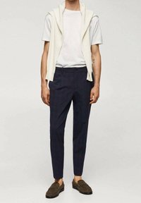 T-shirt blanc, pull crème drapé sur les épaules, pantalon slim-fit bleu marine et mocassins marron foncé avec une finition texturée.
