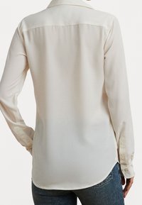 Person i en långärmad, krämfärgad blus med knappade manschetter och blå jeans, sedd bakifrån mot en vit bakgrund.