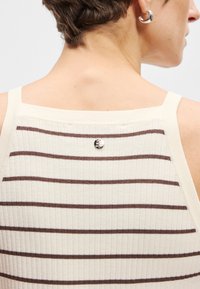 Κρεμ μπλούζα με ραφές, που έχει καφέ οριζόντιες ρίγες, στενές τιράντες και ένα μικρό ασημένιο σήμα στη πίσω πλευρά του λαιμού.