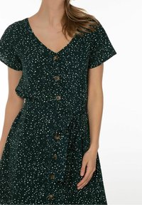 Robe verte à motifs, avec un col en V, des manches courtes, des boutons sur le devant, et un détail de taille nouée avec un design tacheté.