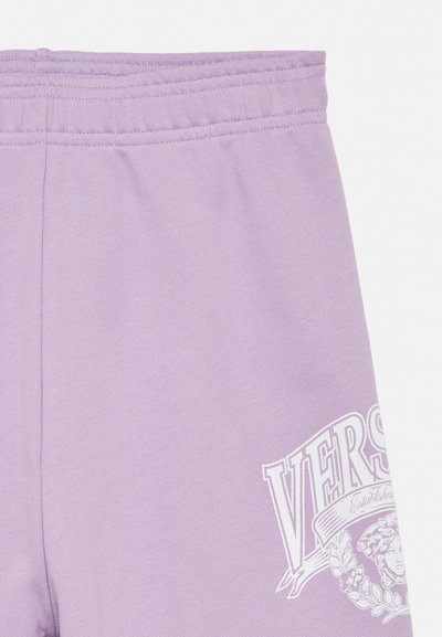 Lila melegítő nadrág elasztikus derékrésszel, részben fehér Versace logóval, melyen Medúza feje és babérkoszorú látható a combon.