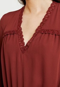 Blusa granate con escote en V, que presenta un borde de encaje a lo largo del escote y pliegues horizontales en la parte inferior. Tejido suave con textura lisa.