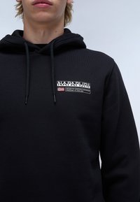 Black hoodie made of soft fabric, featuring a front logo patch with text and flag, drawstrings, and a classic fit.