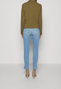 Osoba oblečená v světle modrých džínách, olivově zelené bundě a béžových vysokých kozačkách, stojící proti jednoduchému světlému pozadí.