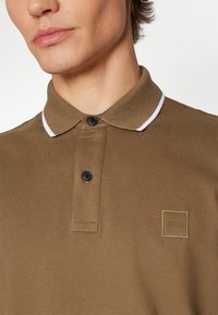 Barna pólóing, texturált anyagból, fehér csíkkal ellátott gallérral, két fekete gombbal és diszkrét márkajelzéssel a mellkasán.
