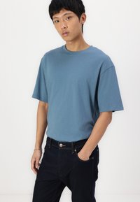 T-shirt de algodão azul com gola redonda e mangas curtas, combinada com jeans de ganga escuros com um bolso frontal e costura visível.