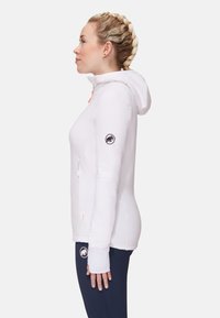 White zip-up hoodie with a textured finish, featured with a hood, hidden pockets, and an embroidered logo on the sleeve. Paired with dark pants.
