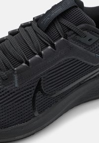 Svart athletic-sko med en teksturert mesh-overdel, kontrasterende glatte detaljer og et fremtredende snøresystem; har en svart swoosh og logodetaljer.