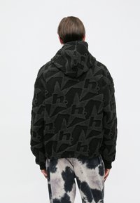 Must kapuutsiga jakk, mis on valmistatud tekstuurse fliisist materjalist ja millel on korduv logomuster. Riietusel on lõdvestunud lõige, koos soonikkoes varrukaotste ja äärega.