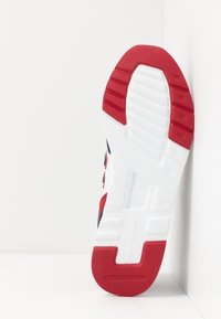 Semelle de basket rouge et blanche en caoutchouc texturé avec des rainures. Présente un logo et un talon incurvé. Motif géométrique visible sous la semelle.