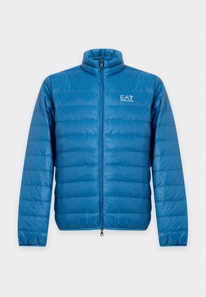 Homme portant une doudoune bleue, un bonnet noir, une chemise bleu marine, un pantalon beige et des baskets noires et blanches, debout avec une main dans la poche.