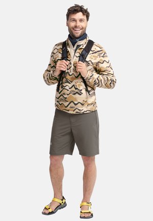 Lachende man met een patroonfleece, donkere korte broek, gele sandalen en zwarte rugzakriemen, staand tegen een witte achtergrond.