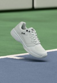 Λευκό αθλητικό παπούτσι FILA που αιωρείται πάνω από μια μπλε και πράσινη επιφάνεια τένις, κοντά σε μια λευκή γραμμή ορίου.