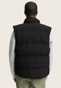 Zwarte gewatteerde vest met een hoge kraag, met horizontaal gequiltde secties. Draagt over een grijze longsleeve shirt, met een effen achtergrond.