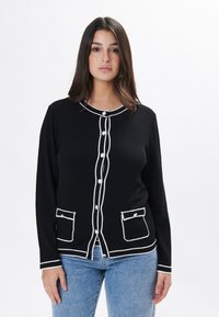 Cárdigan negro con ribete blanco, con cuello redondo, cierres de botones y dos bolsillos frontales. Fabricado en suave tejido de punto.