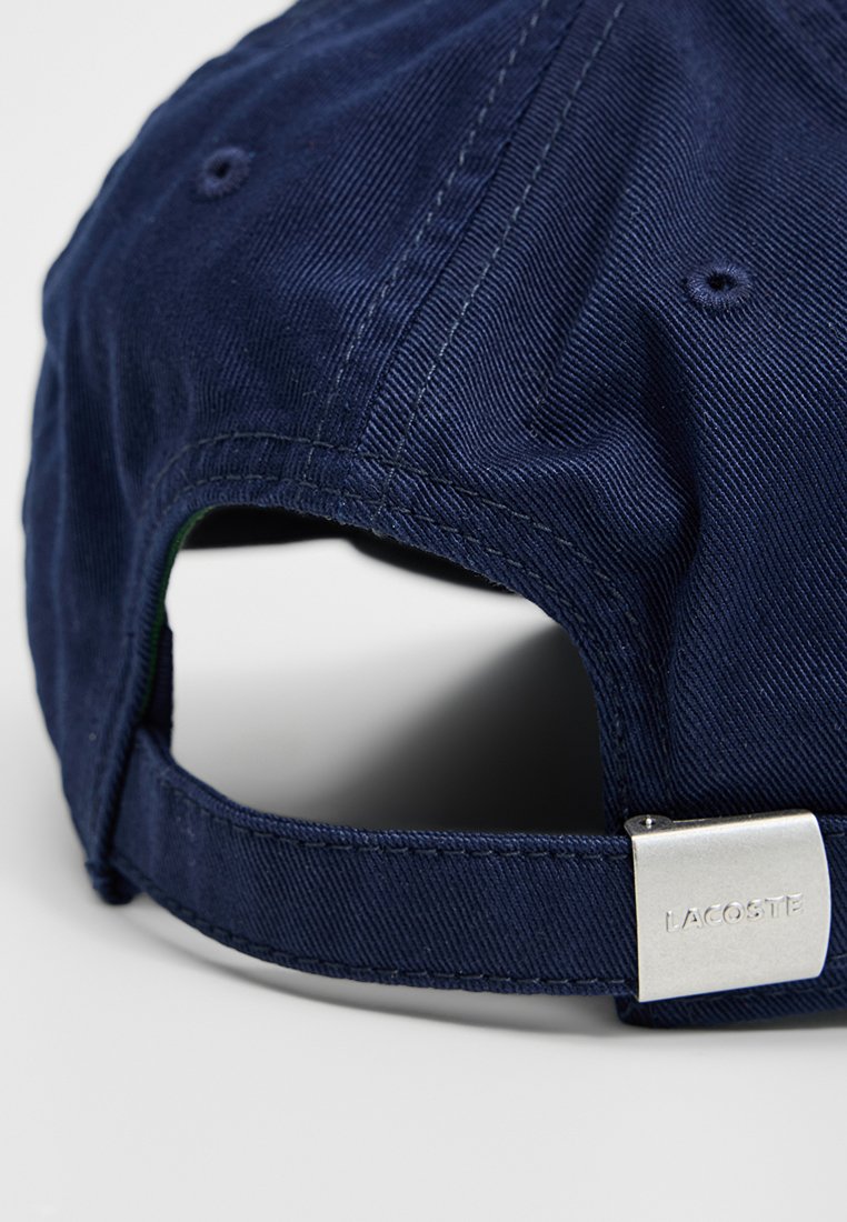 Gorra de color azul marino hecha de sarga de algodón con una correa ajustable. Presenta un acento metálico plateado con "LACOSTE" grabado. Superficie texturada.