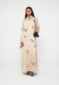 Hosszú bézs ruha virágos hímzéssel, gallérral, kulcslyuk nyakkivágással és hosszú ujjal. A modell egy kis fekete táskát tart.