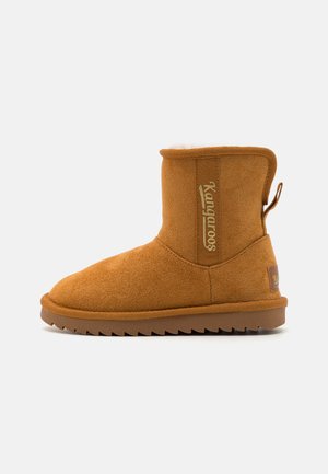 Tanhoge suède enkellaars met een zachte voering, een gestructureerde geelbruine buitenkant, een platte zool en een logo-detail aan de zijkant en achterkant.