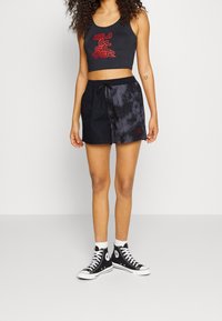 Černý crop top s červeným logem, kombinovaný s černými šortkami s batikovaným vzorem a černými vysokými teniskami s bílými detaily. Neformální sportovní souprava.