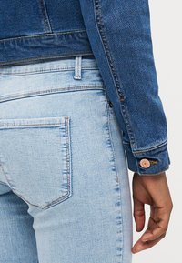Denim jakna v srednje modrem odtenku s rahlo zbledelim videzom, v kombinaciji s svetlo modrimi kavbojkami, ki imajo zadnje žepe in detajle šivanja.