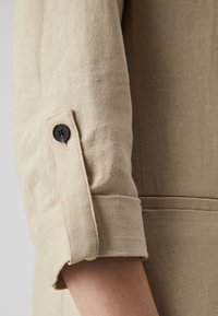 Beige liinakangas hiha, rullattu ylös ja kiinnitetty mustalla nappiläpällä, mikä paljastaa osan kyynärvarresta ja vaatteen sivusauman.