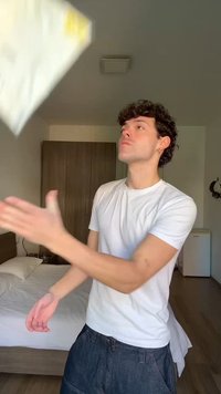 Junger Mann mit weißem T-Shirt wirft ein Stück Papier in einem Schlafzimmer mit kleinem Holzkleiderschrank, Bett und kleinem Kühlschrank im Hintergrund.