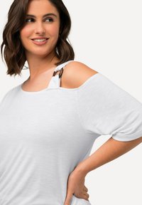 Camiseta blanca de gran tamaño con escote redondeado y cortes en los hombros, que presenta una tela texturizada y detalles de tirantes ajustables.