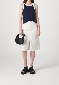 Débardeur bleu marine avec bordure blanche, jupe blanche jusqu'aux genoux avec une fente à l'avant, et sac à main noir. Des sandales noires épaisses complètent la tenue.