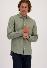 Man past een manchette van een groene herringbone-patroon lange mouw shirt, draagt donkerblauwe jeans en staat tegen een effen lichte achtergrond.