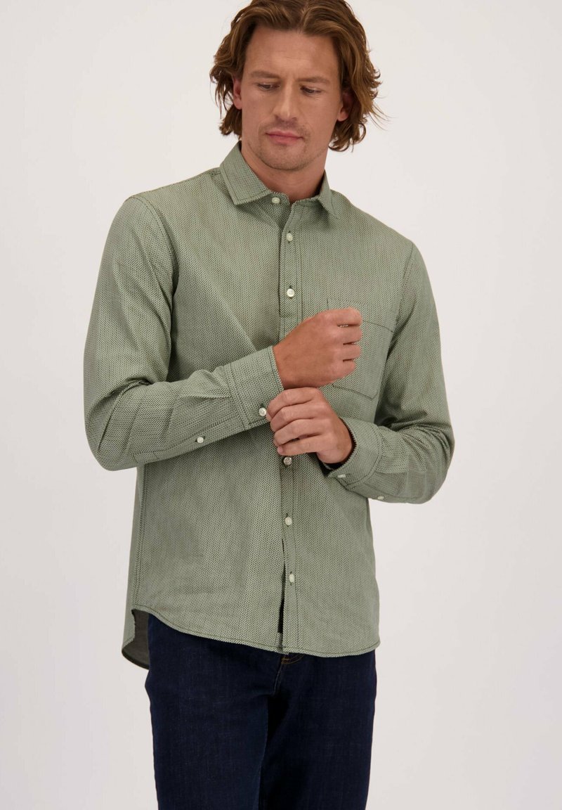 Man past een manchette van een groene herringbone-patroon lange mouw shirt, draagt donkerblauwe jeans en staat tegen een effen lichte achtergrond.