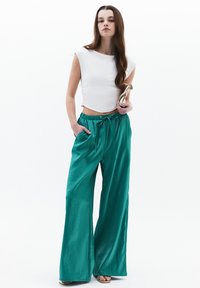 Witte cropped top en teal wijde broek met een elastische tailleband en zakken. Model houdt een gouden clutch vast. Gladde, glanzende textuur.
