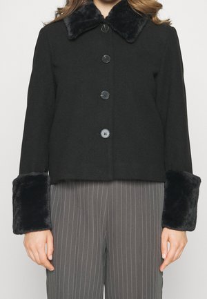 Chaqueta negra con botones y cuello y puños de piel sintética, usada sobre pantalones gris con rayas finas.