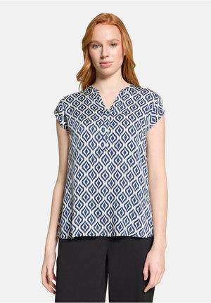 Junge Frau mit roten Haaren, die eine kurzärmelige blau-weiße Bluse mit geometrischem Muster und schwarze Hose trägt, steht vor einem einfarbigen Hintergrund.