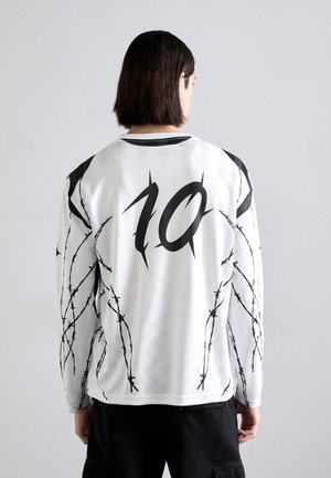 Maillot à manches longues blanc avec des graphismes de fil barbelé noir. Présente un grand numéro noir 10 au dos, terminé par un col noir.