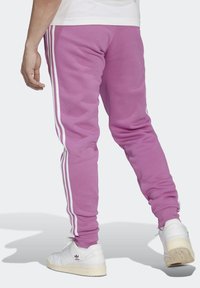 Lila Jogginghosen mit weißen Seitenstreifen, einer schmal zulaufenden Passform und einer weichen Textur, kombiniert mit weißen Sportschuhen, die ein Logo zeigen.