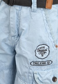 Pantaloni corti azzurri realizzati in tessuto morbido, con una tasca visibile, dettagli in pelle marrone e loghi ricamati in filo scuro.