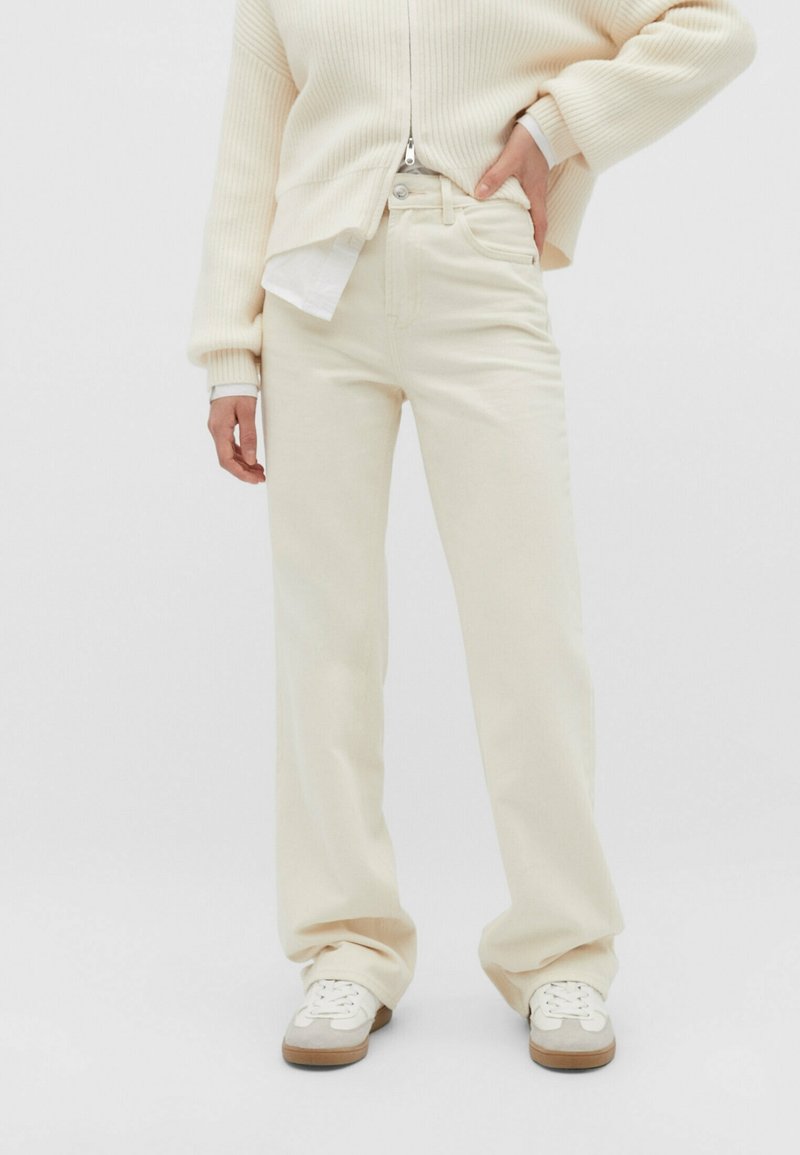 Personne portant un jean droit couleur crème, un pull à fermeture éclair crème et des baskets blanches sur un fond uni, main posée sur la hanche.