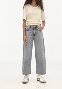 Jeans grigi con gamba larga e vita alta, abbinati a una blusa leggera color crema. Le scarpe includono sneakers bianche con dettagli grigi.