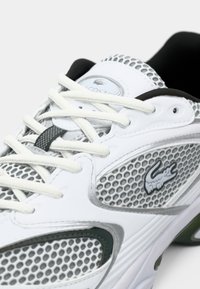 Lacoste sneaker in white with mesh panels, round laces, gray accents, logo detail, and cushioned sole for breathability and comfort.