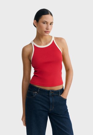 Femme aux cheveux foncés attachés en chignon, portant un haut rouge sans manches avec des bordures blanches et un jean bleu foncé, debout avec une main dans la poche.