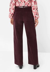 Pantalon large en velours côtelé bordeaux avec deux poches arrière, tissu texturé et taille haute, associé à un chemisier à motifs et des bottes noires à talons.