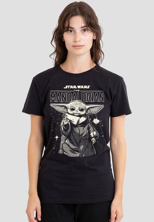 Junge Frau trägt ein schwarzes Star Wars The Mandalorian T-Shirt mit einer detaillierten Grafik von Baby Yoda vor einem sternenklaren Hintergrund.