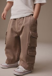 Bruine cargobroek met meerdere zijzakken, middellicht gewicht stof en losse pasvorm, gecombineerd met witte sneakers met rode strepen.