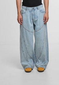 Helle blaue, weit geschnittene Jeans mit abgerundeten Nahtdetails, vorderen Taschen und Knopfverschluss. Kombiniert mit gelben Schuhen.