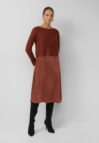 Rostfarbener Strickpullover über einem Kleid mit satinähnlichem Finish in derselben Farbe, kombiniert mit knielangen schwarzen Wildlederstiefeln. Glatte Textur, lange Ärmel.