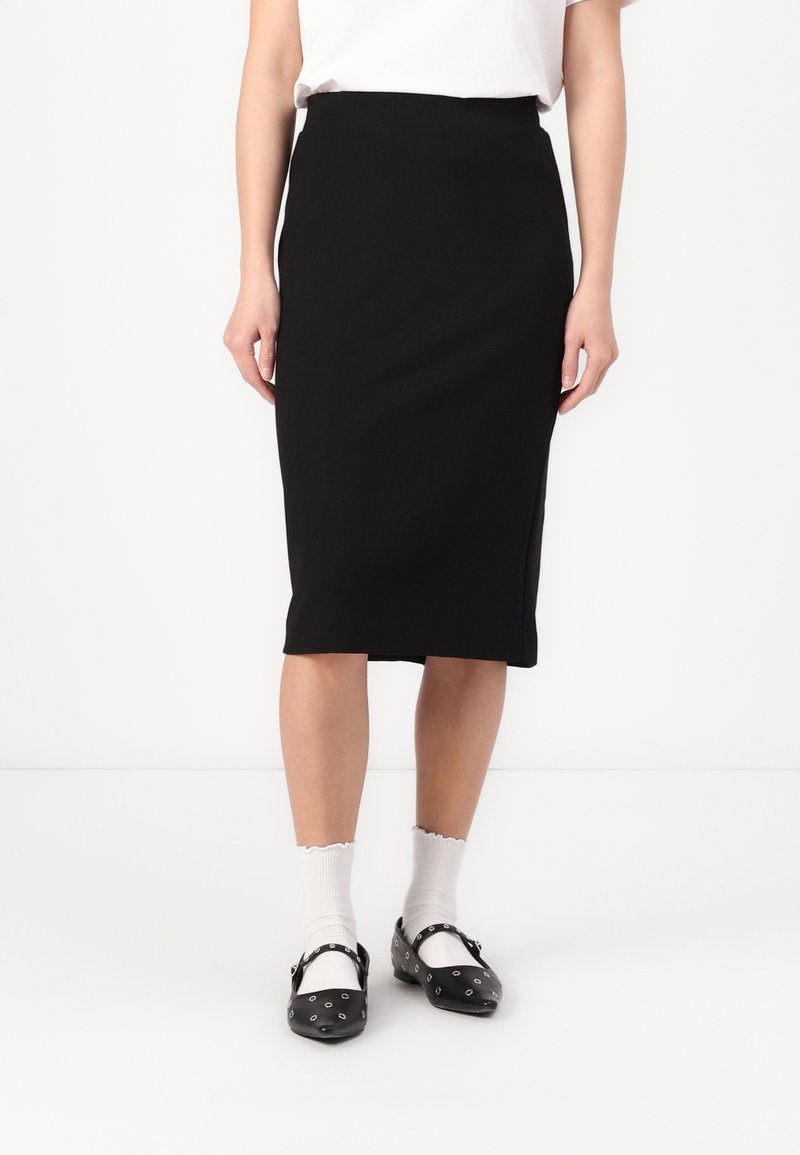 Falda lápiz negra hasta la rodilla con una tela suave y elástica y una silueta ajustada, combinada con zapatos negros con acentos metálicos.