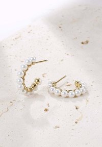 Pendientes de aro dorados con perlas falsas blancas, diseño circular con cuentas de diferentes tamaños, sobre un fondo texturizado y claro.