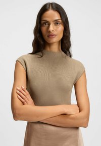 Woman with long dark hair wearing a sleeveless beige knit top, arms crossed, against a plain white background.