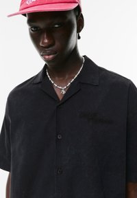 Cămașă scurtă neagră cu mâneci scurte, fabricată dintr-un material texturat, având un logo brodat "Out of The Norm". Este purtată cu un colier argintiu și o șapcă roz.